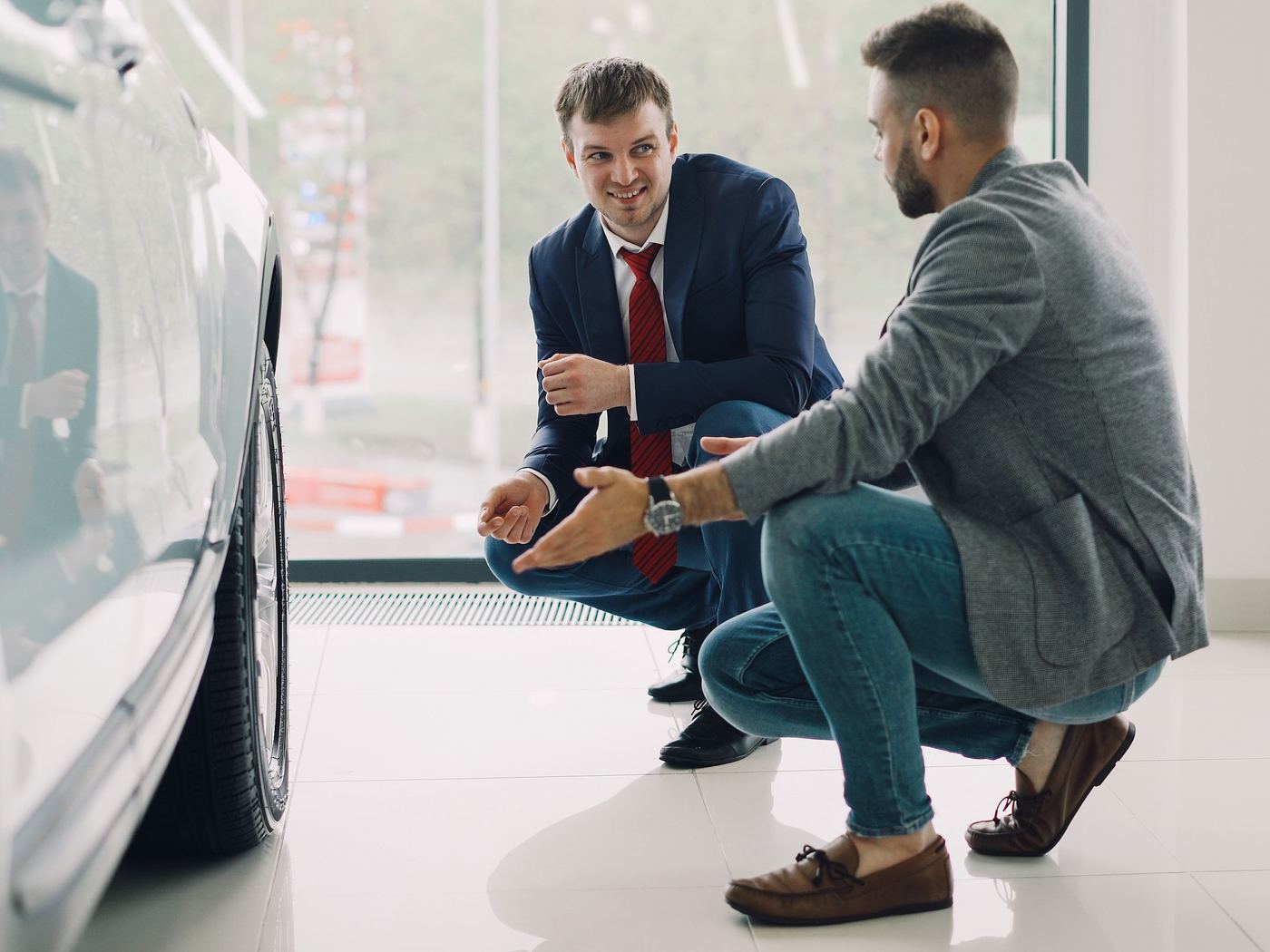 A salesperson and a customer crouched beside a car on a showroom floor, mid-conversation.